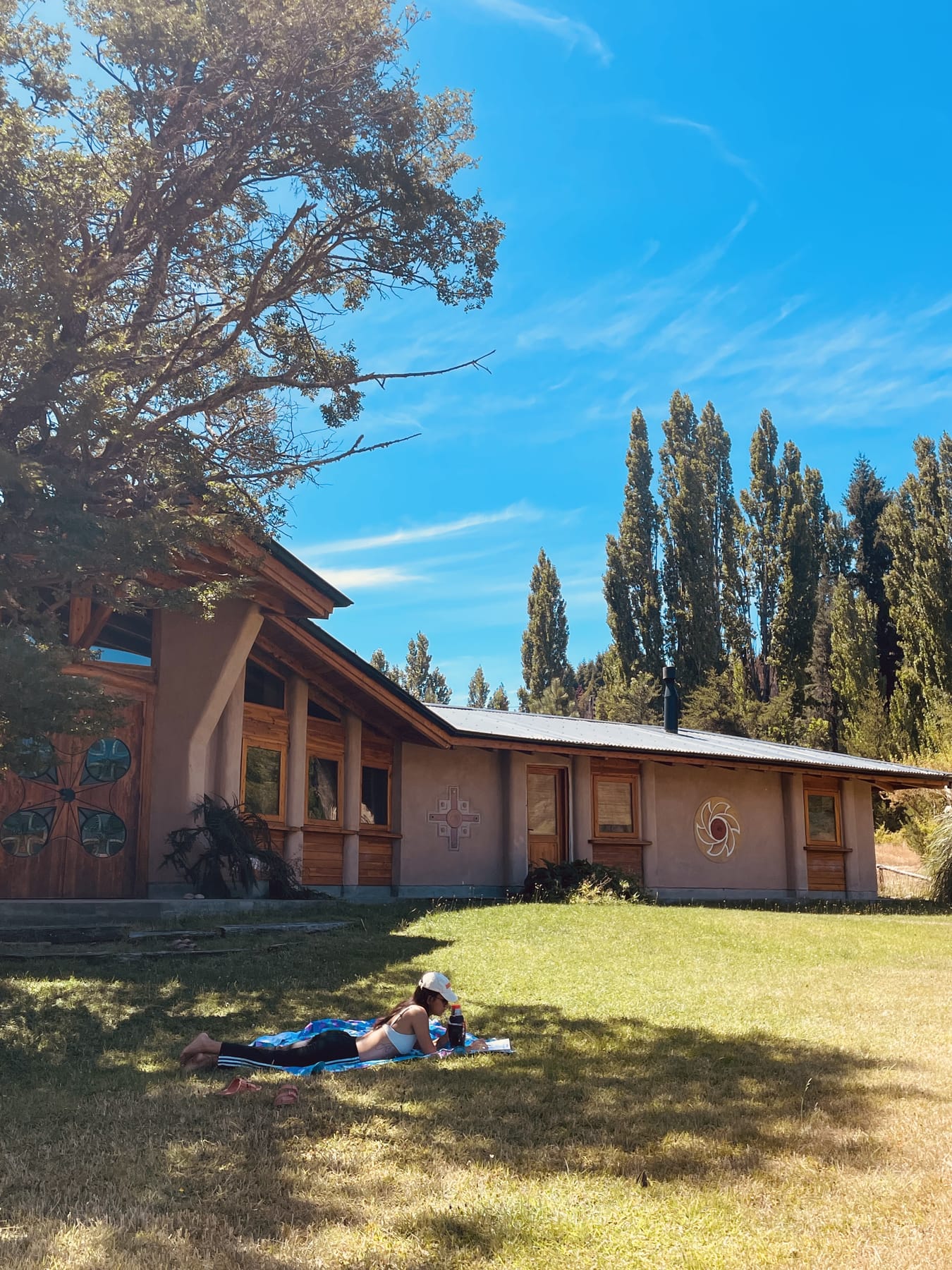 La casa de La Tregua, con mandalas en sus paredes, jardín, un gran árbol y álamos bajo el cielo azul de El Bolsón
