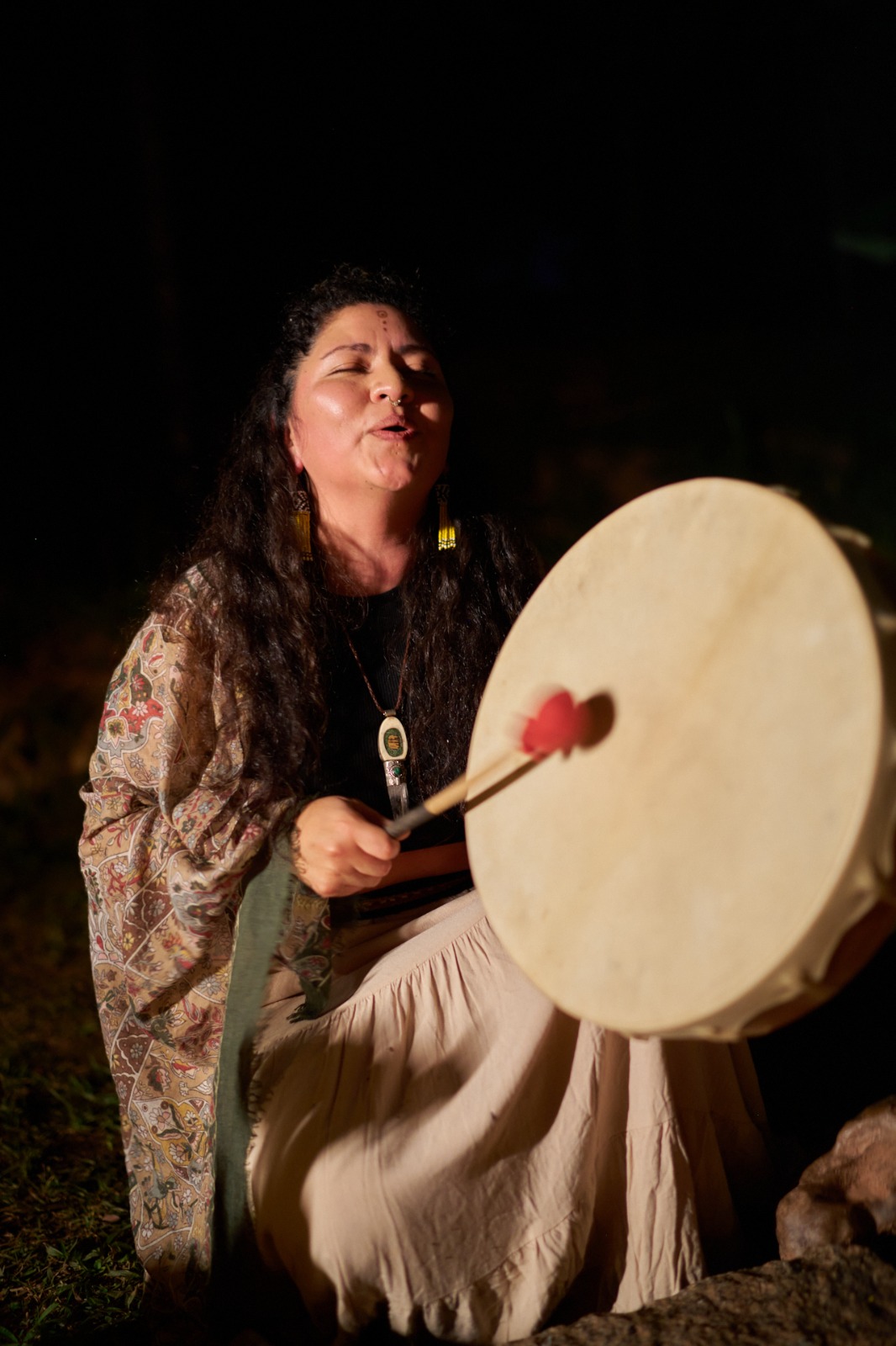 Facilitadora cantando y tocando un tambor chamánico de noche, junto al fuego